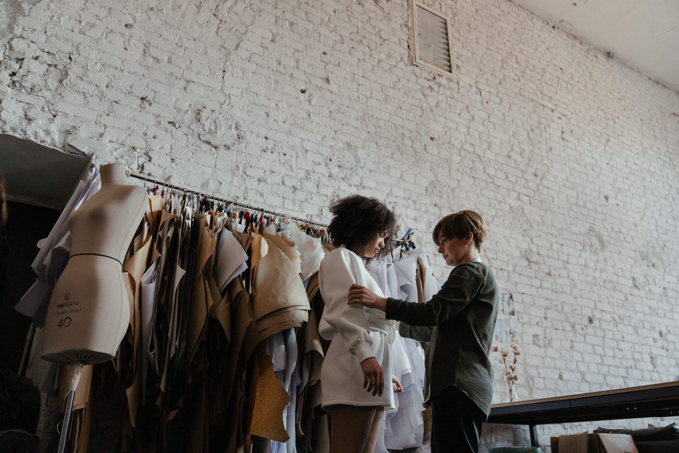 Two designers fitting a white jacket beside a rail of patterns and dress forms
